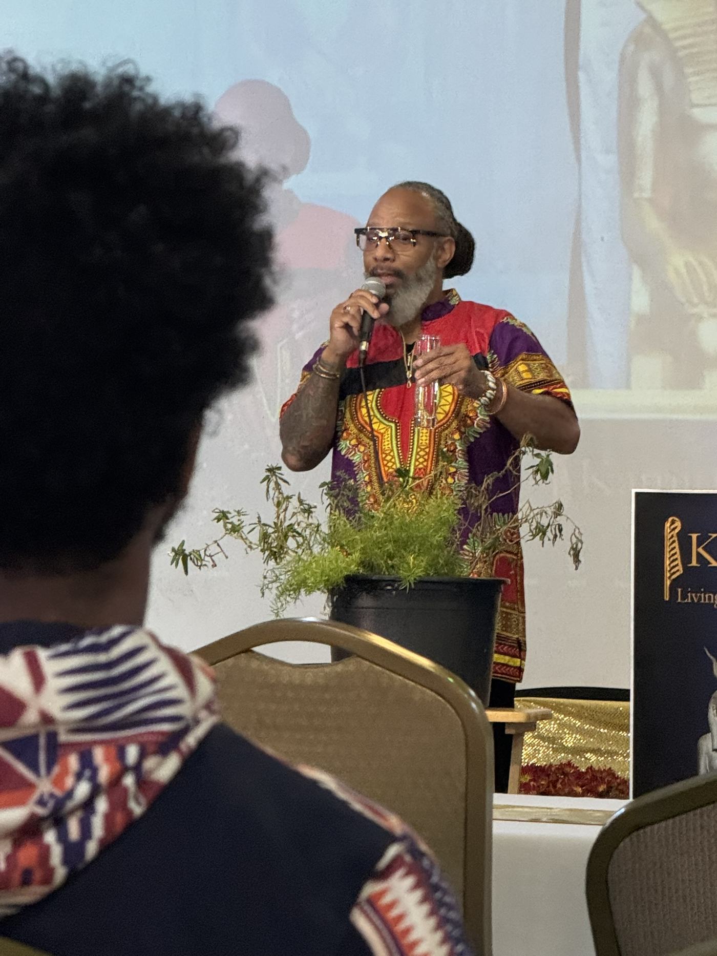 Heru TchaasAmen speaking at a teaching event with Kamitic imagery projected behind him